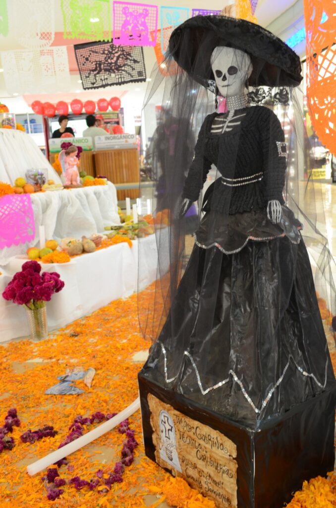 Figura de la Catrina con sombrero y vestido negro sobre una ofrenda de cempasúchil